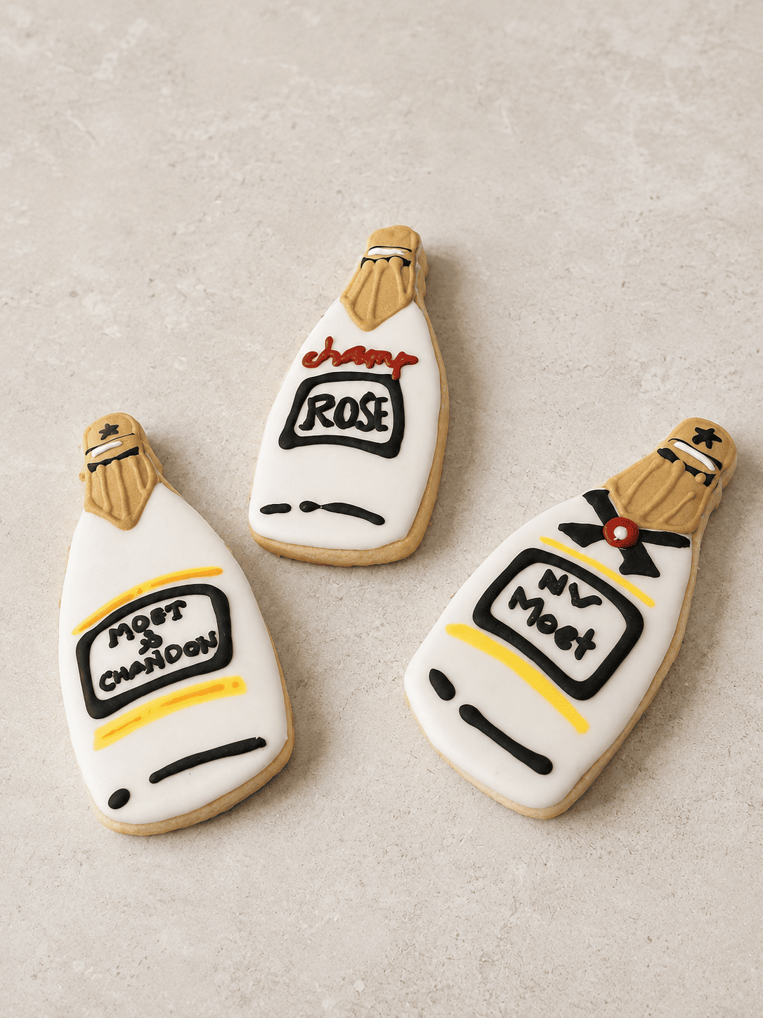 Three champagne-bottle sugar cookies iced in white, gold, and black on a stone surface